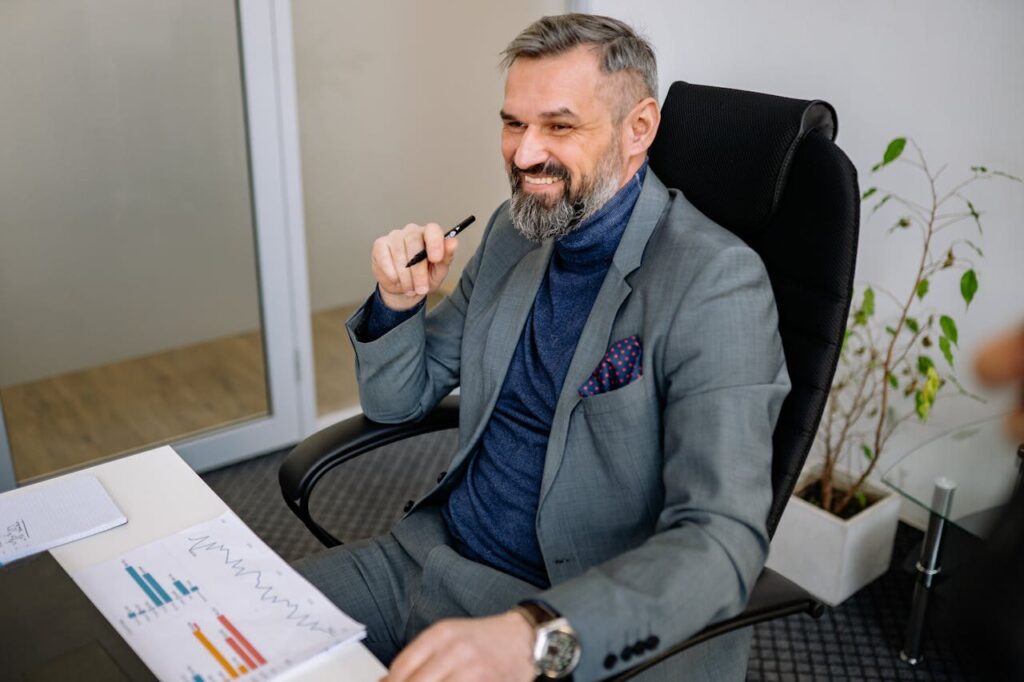 pexels photo 7792844 Smiling businessman in a suit sits at a desk, illustrating office professionalism.